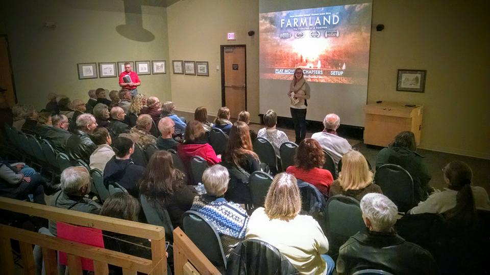 An audience sits in a room facing a screen displaying the title screen for the documentary “Farmland,” with a speaker standing at the front.