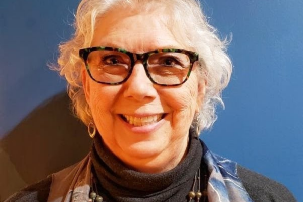An older woman with short gray hair and glasses smiles at the camera. She is wearing a dark turtleneck, a beaded necklace, and a scarf, standing in front of a blue wall.