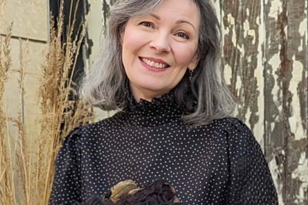 A woman with shoulder-length gray hair smiles, wearing a black polka dot blouse. She stands against a textured wooden background with dried plants on the side. She holds a bundle of dark fabric or material in front of her.