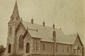 Sepia-toned historical photograph of a stone church with a tall steeple, arched windows, and ornate roof details, set on bare ground with no visible surrounding buildings.