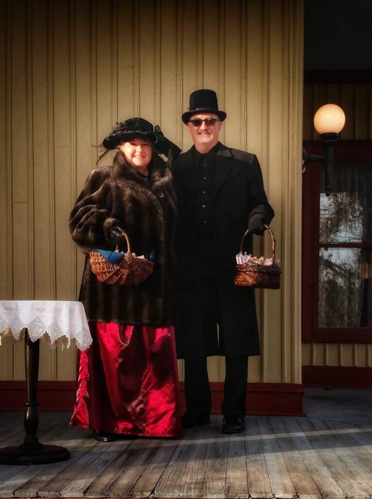 Color photo of a man and woman dressed in outdoor Victorian garb handing out candy from wicker basekts to trick-or-treaters