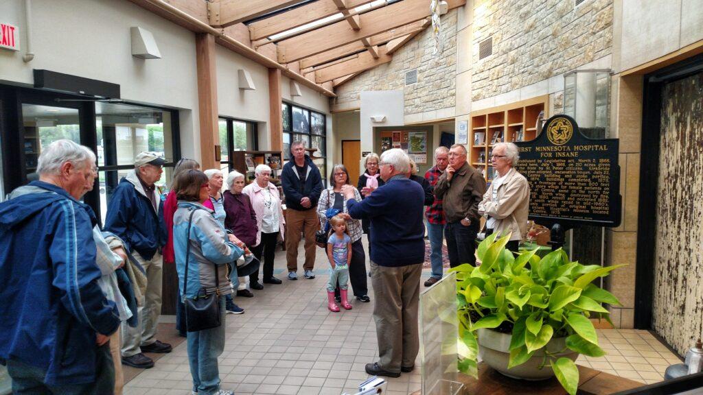 Color photo of a male tour guide speaking to and in front of a group of senior citizens in the lobby of a museum