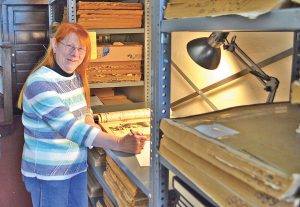 Color photo of a redheaded adult female standing in front of pallet racking filled with old newspaper ledgers