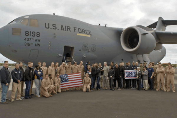 Color photograph of a group of people standing in front of a military airplane. One group of people is holding a U.S. flag. Another group is holding a USO sign.