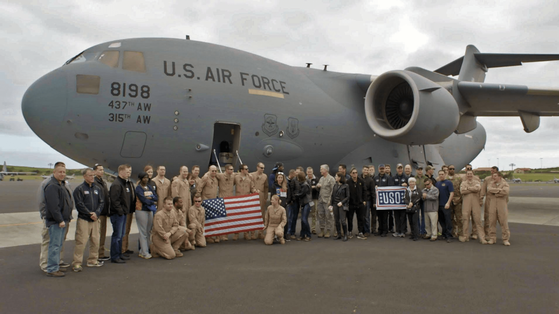 Color photograph of a group of people standing in front of a military airplane. One group of people is holding a U.S. flag. Another group is holding a USO sign.