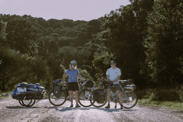 Color photo of two adults standing in the middle of a paved road with green trees in the background. Male and female are both standing in front of bicycles.