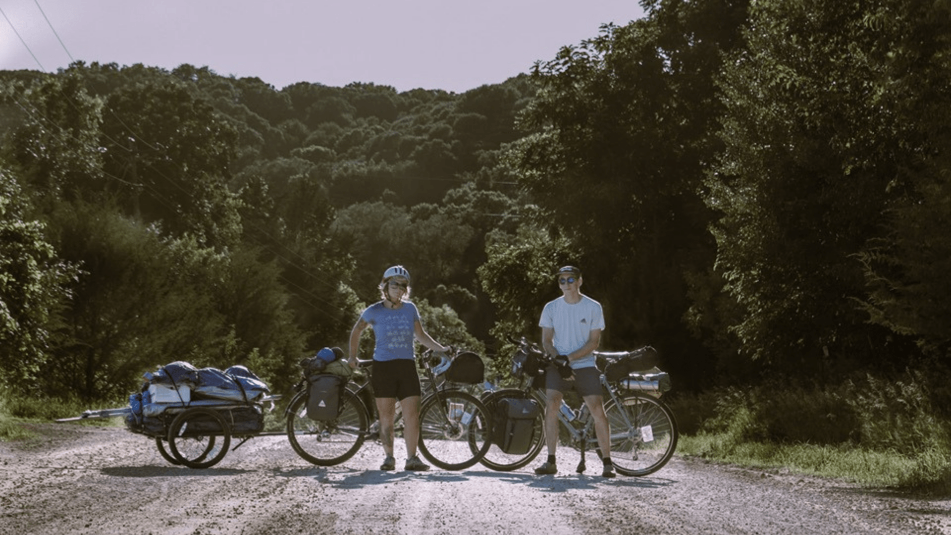 Color photo of two adults standing in the middle of a paved road with green trees in the background. Male and female are both standing in front of bicycles.