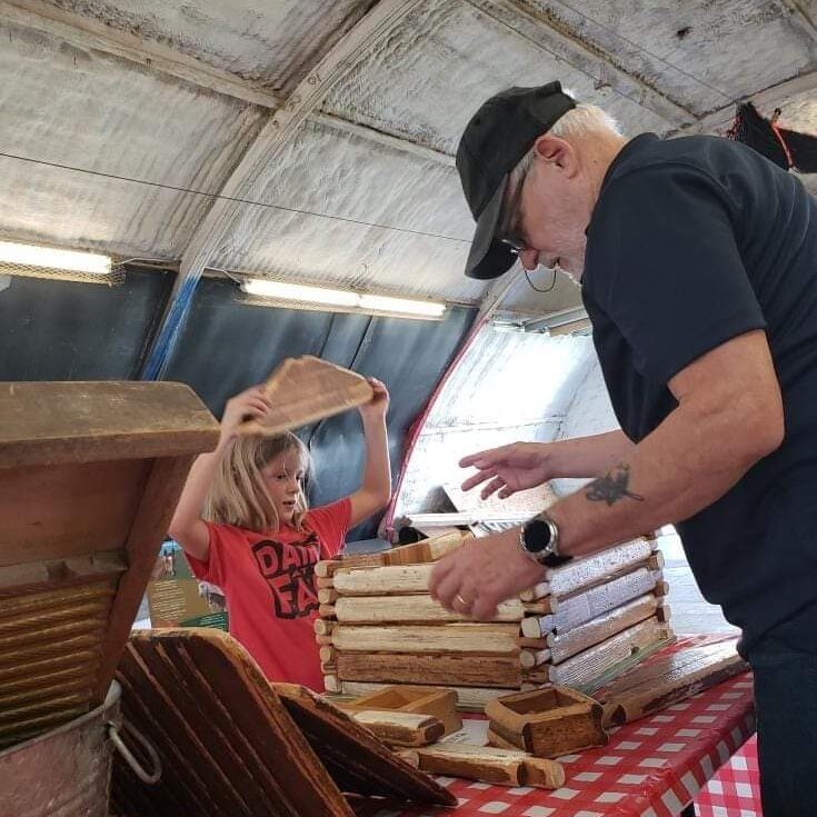 An older man and a young child build a structure with log-like pieces on a table covered in a red and white checkered cloth. The child holds a piece of wood above their head. The setting appears to be an indoor, rustic location.