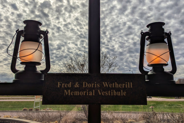 Two lanterns are mounted on either side of a sign reading “Fred & Doris Wetherill Memorial Vestibule,” with a cloudy sky and grassy area visible in the background.