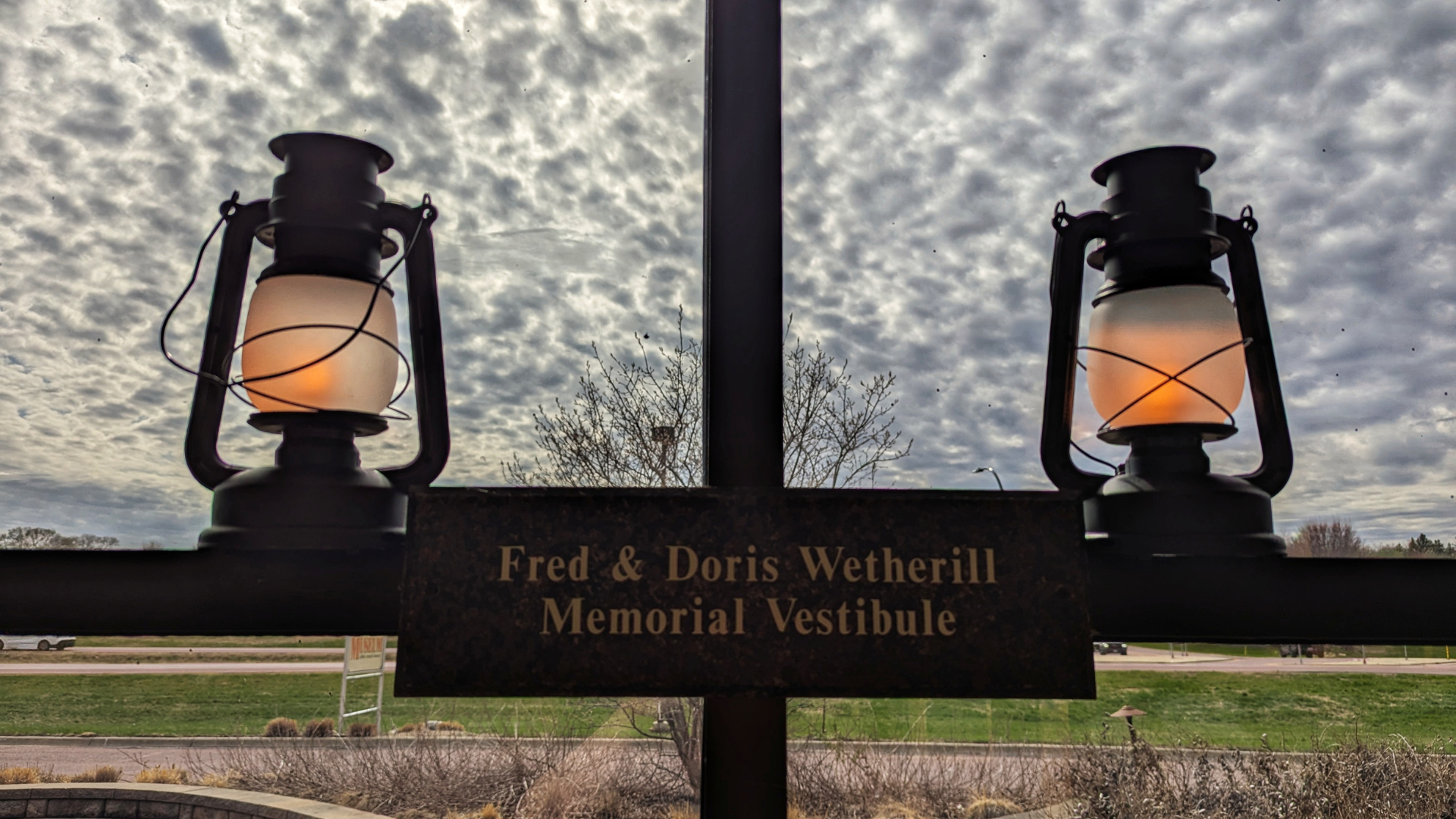 Two lanterns are mounted on either side of a sign reading “Fred & Doris Wetherill Memorial Vestibule,” with a cloudy sky and grassy area visible in the background.