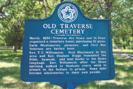 A blue historical marker sign for Old Traverse Cemetery describes its founding in 1856, burial of missionaries, pioneers, and Civil War veterans, and efforts to minister and educate the Sioux people. Trees and grass are visible behind.