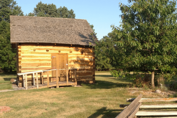 A small wooden cabin with a shingled roof and a ramp stands on a grassy area, surrounded by trees. A wooden fence and a tree are visible in the foreground on a sunny day.
