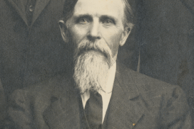 An older man with a long white beard and mustache, wearing a dark suit and tie, is seated and looking at the camera. The background is partially visible, showing parts of other people in similar attire.