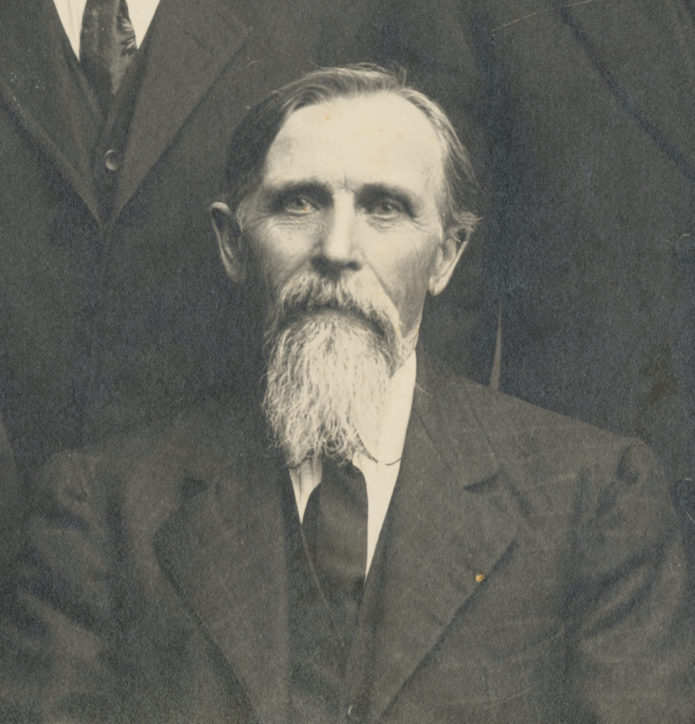 An older man with a long white beard and mustache, wearing a dark suit and tie, is seated and looking at the camera. The background is partially visible, showing parts of other people in similar attire.
