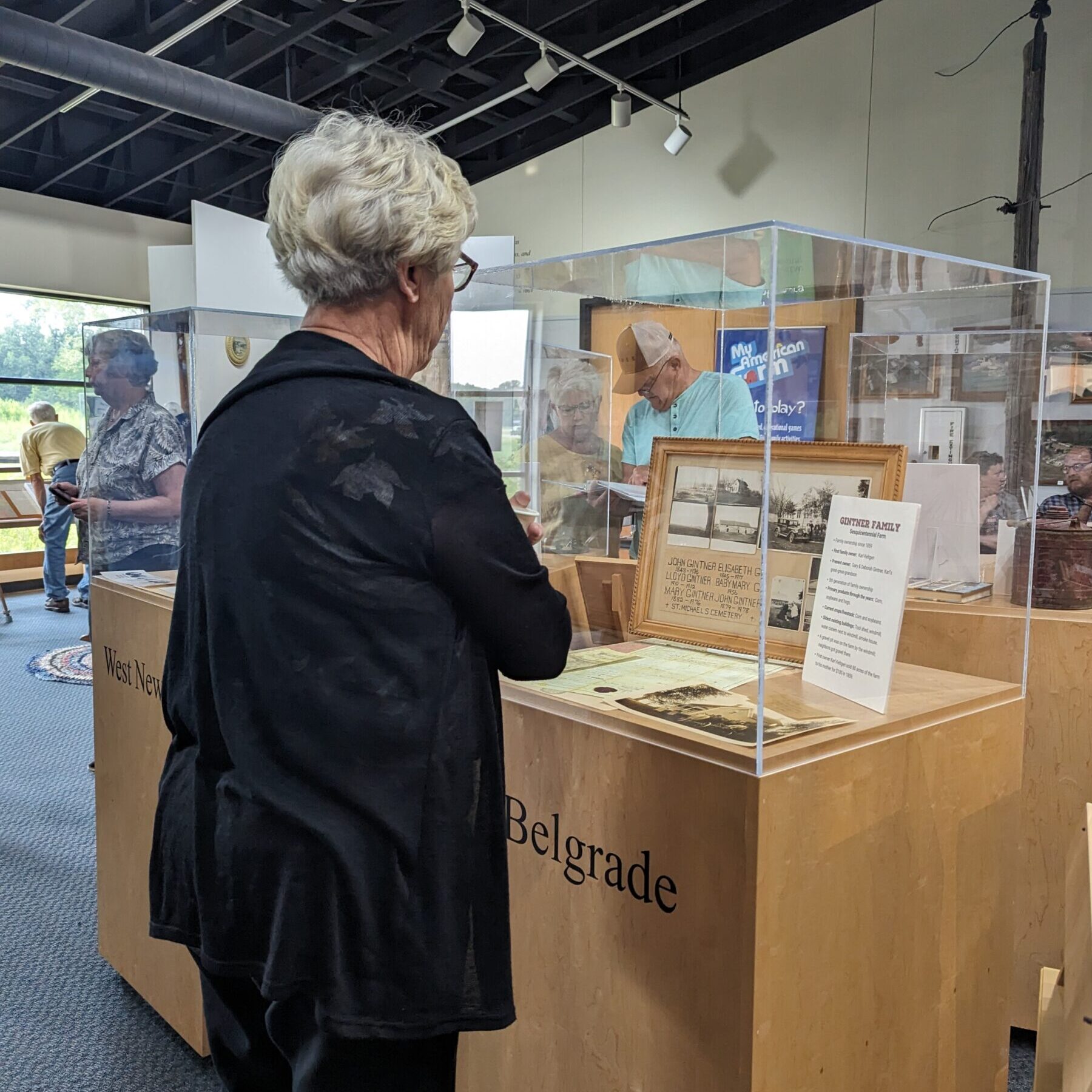An older woman stands in front of a glass display case labeled "Belgrade" at a museum exhibit, looking at historical documents and photographs; other people view exhibits in the background.