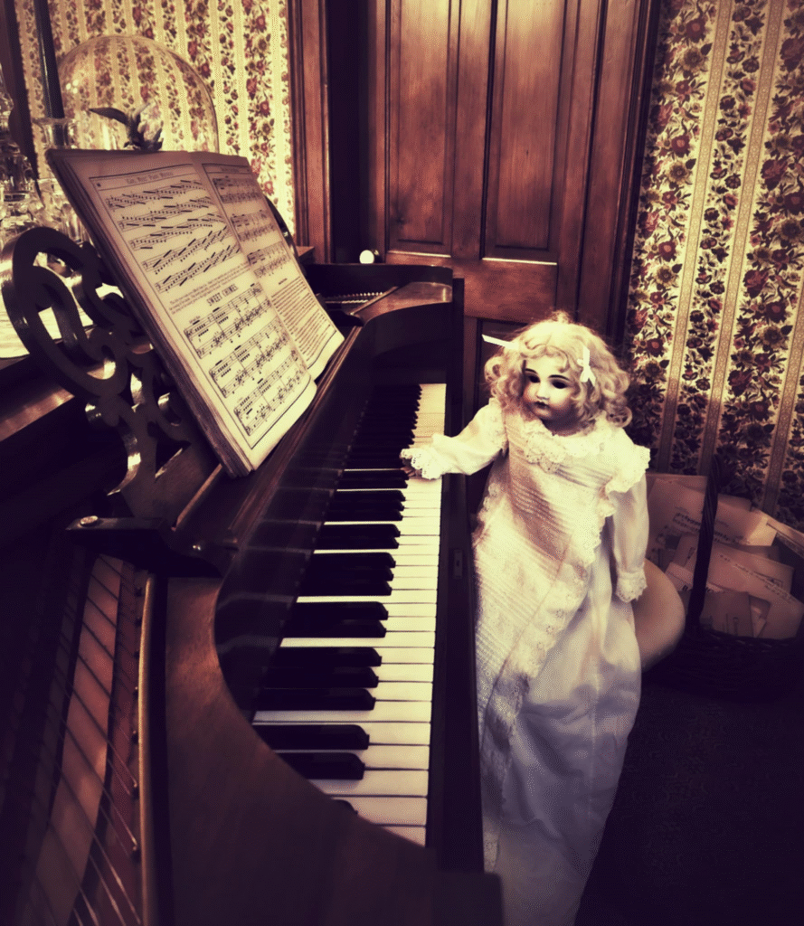 A porcelain doll in a white dress stands next to an upright piano with sheet music on its stand. The scene is set in a room with floral wallpaper and a wooden door in the background.