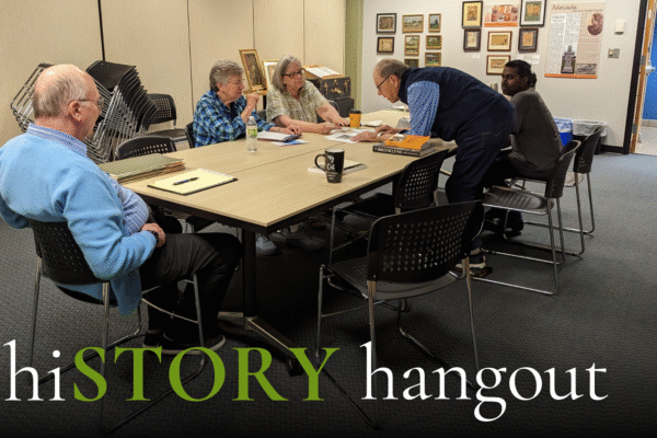 Five people sit around a table in a room with beige walls, looking at papers and books. Framed photos and documents are displayed on the back wall. The text "hiSTORY hangout" appears at the bottom.