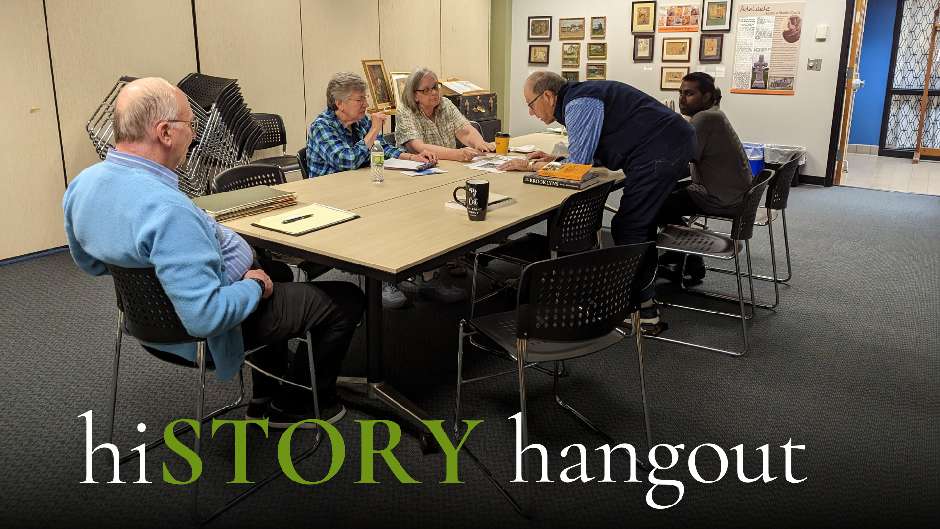 Five people sit around a table in a room with beige walls, looking at papers and books. Framed photos and documents are displayed on the back wall. The text "hiSTORY hangout" appears at the bottom.