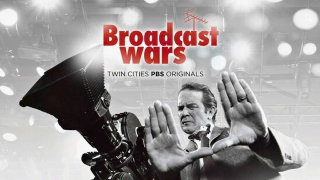 A man stands behind a large studio camera, framing a shot with his hands. The text "Broadcast Wars, Twin Cities PBS Originals" appears above him on a background of lights and equipment.