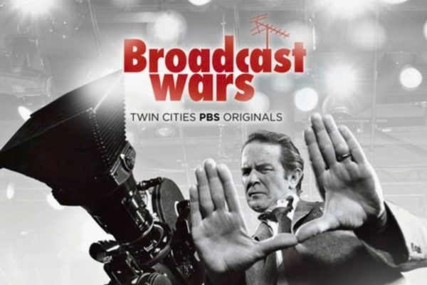 A man stands behind a large studio camera, framing a shot with his hands. The text "Broadcast Wars, Twin Cities PBS Originals" appears above him on a background of lights and equipment.