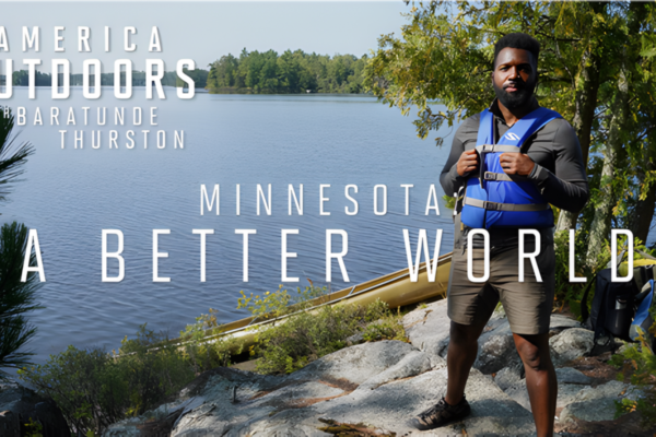 A man wearing a blue life vest stands on a rocky shore next to a lake with trees in the background. Text on the image reads: "America Outdoors with Baratunde Thurston. Minnesota: A Better World.