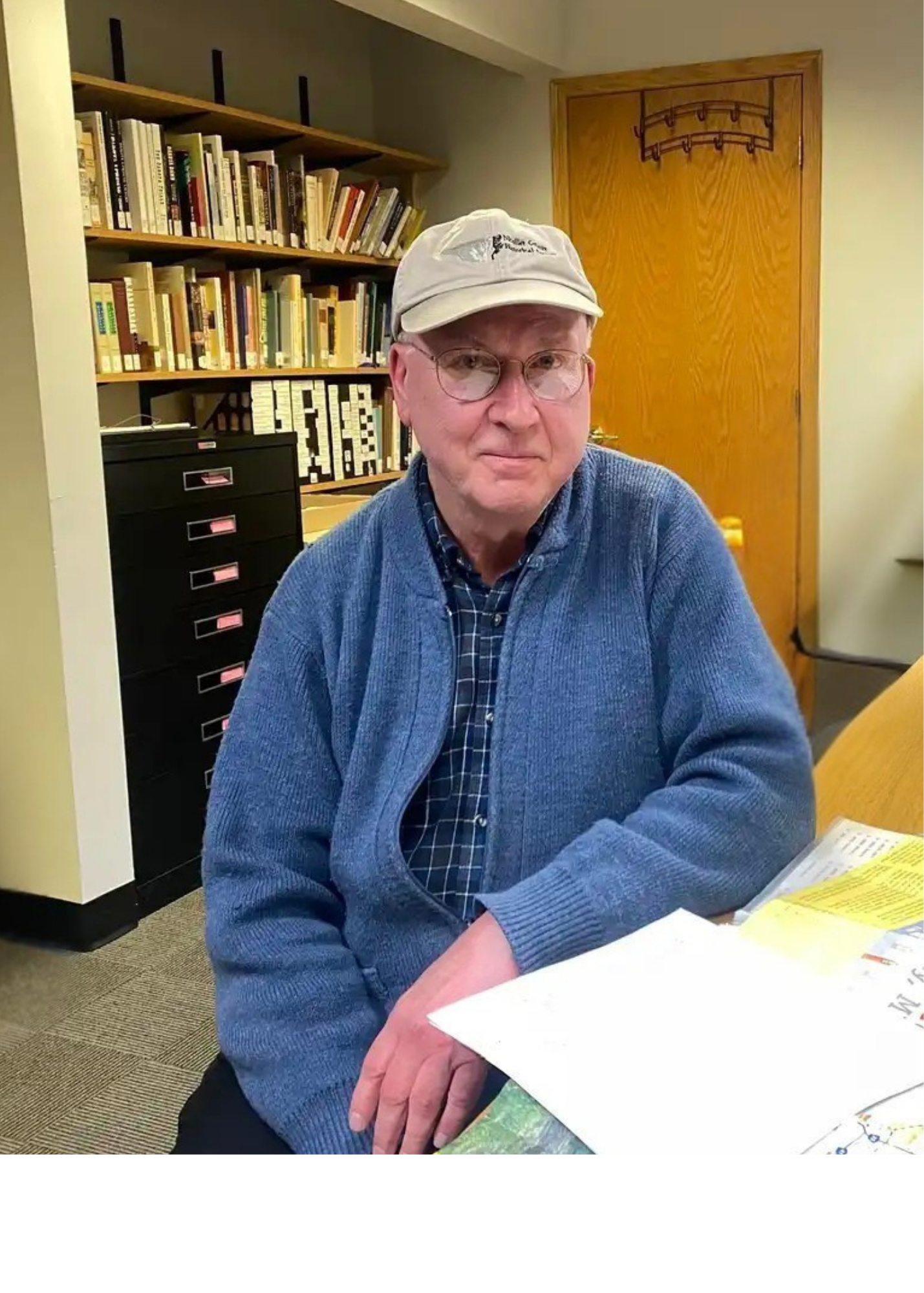 An older man wearing glasses, a light gray cap, and a blue sweater sits at a desk with papers in front of him. Behind him are bookshelves filled with books and files, and a closed wooden door.