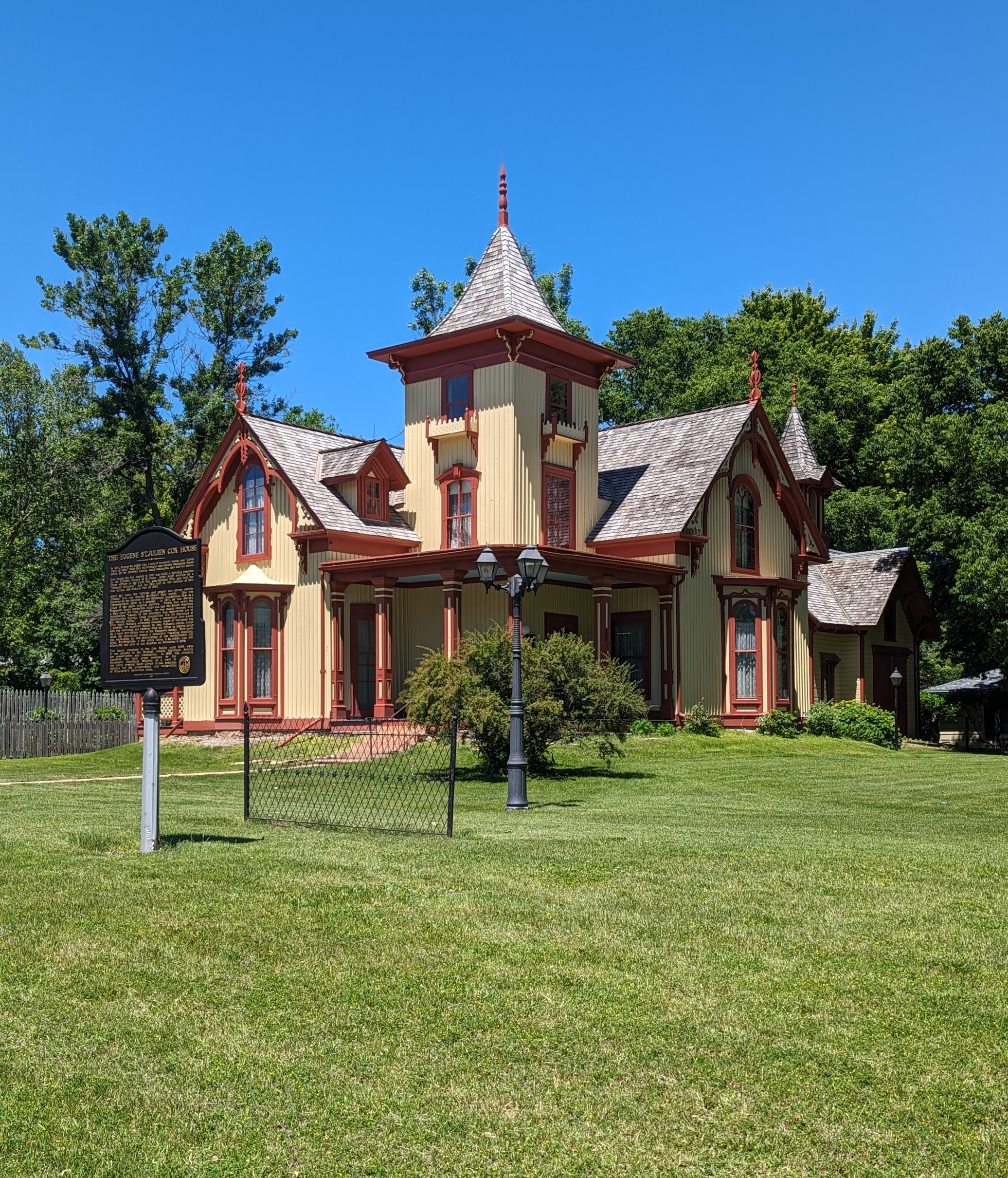A large, pale yellow Victorian-style house with red trim, steep gabled roofs, and a central tower stands on a green lawn. A historical marker hints at the Cox House self-guided tour; a black fence, trees, and blue sky complete the scene.