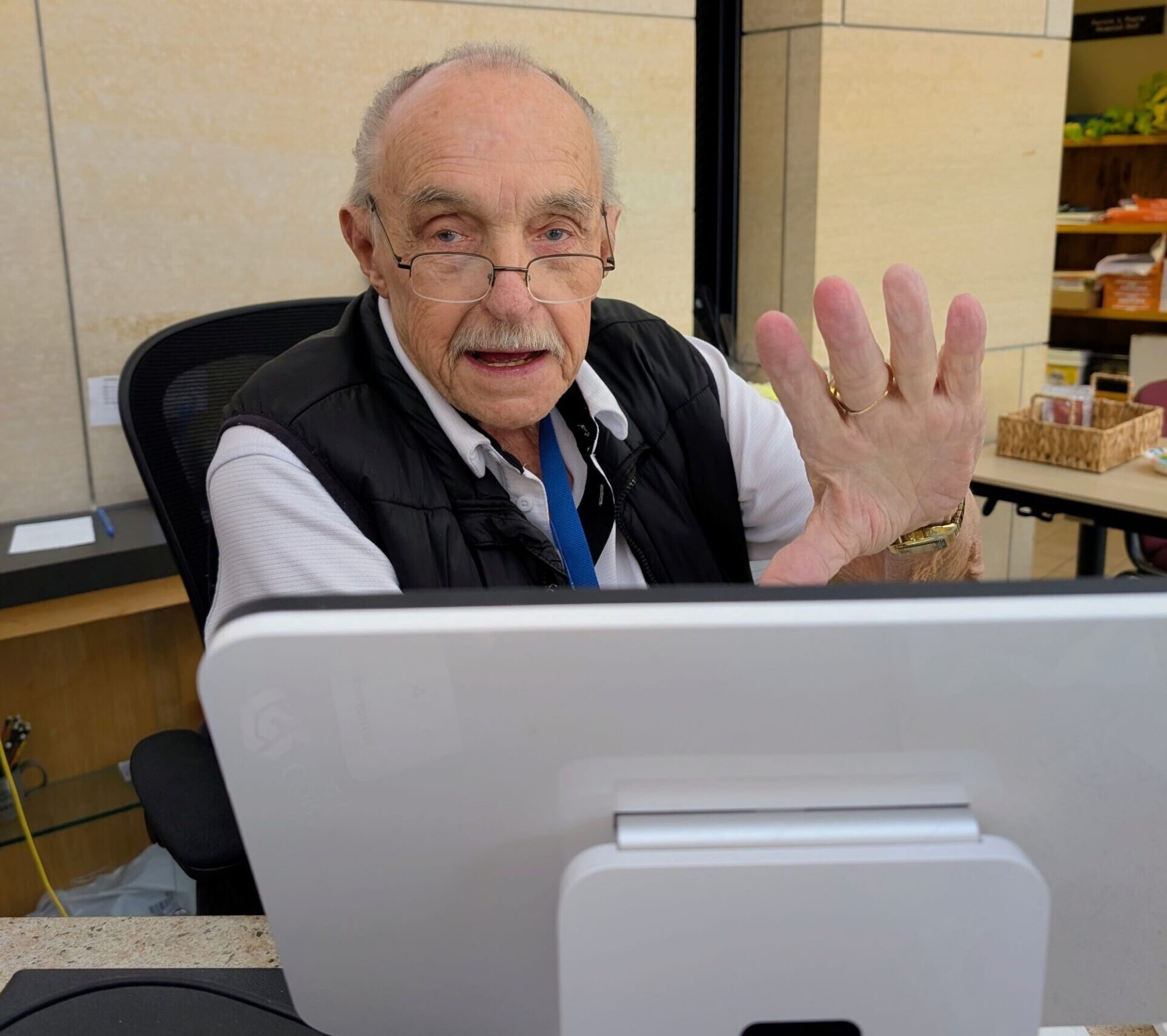 An older man with glasses and a mustache sits behind a counter, looking at the camera and raising his hand in greeting. He is in front of a computer terminal, with a sign and shelves in the background.