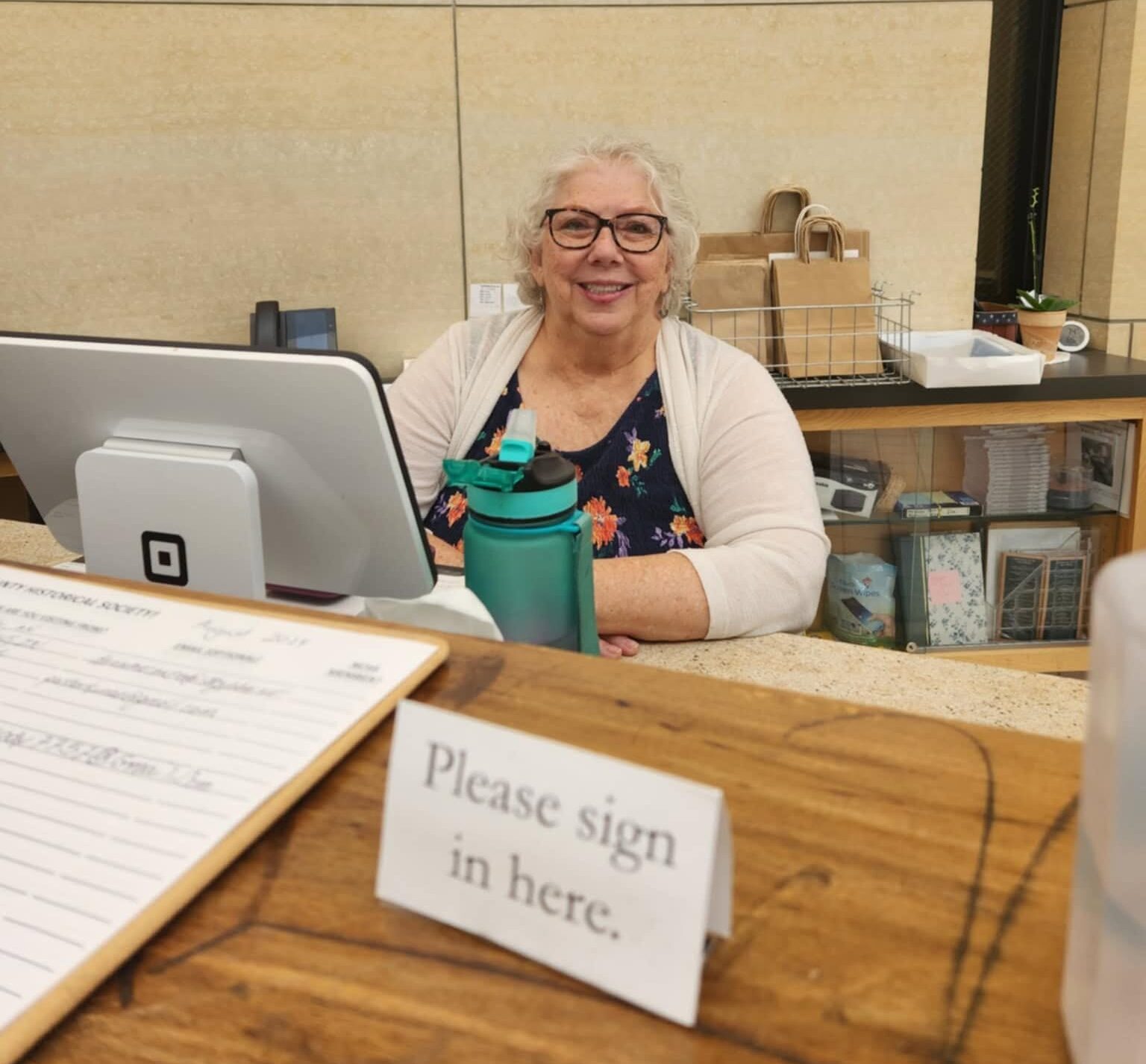 An older man with glasses and a mustache sits behind a counter, looking at the camera and raising his hand in greeting. He is in front of a computer terminal, with a sign and shelves in the background.