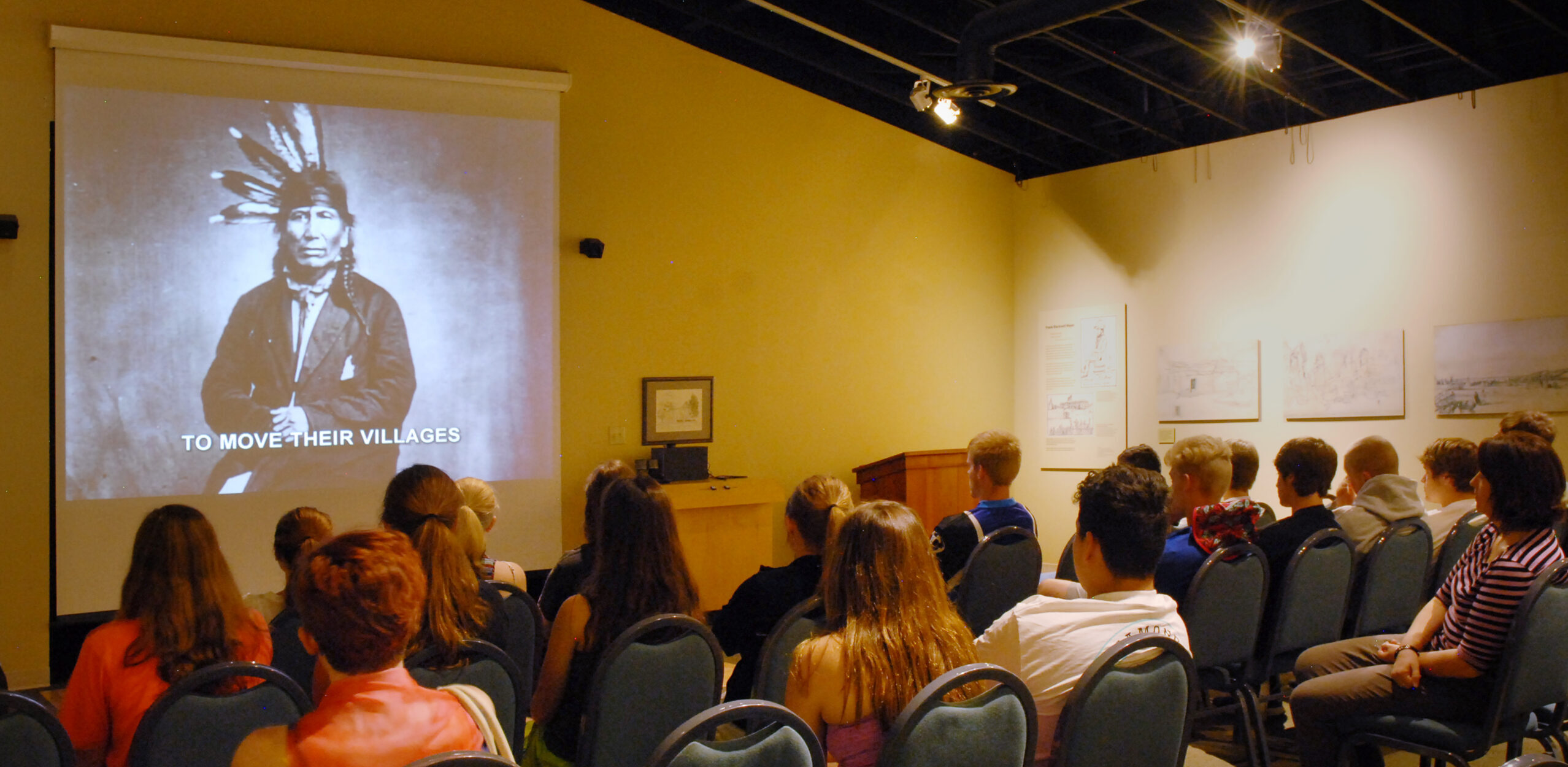 A group of people sit in rows of chairs at the Nicollet County Historical Society Treaty Site History Center, watching a projected black-and-white image of a person in a feathered headdress with the text “TO MOVE THEIR VILLAGES” in a dimly lit room adorned with art on the walls.