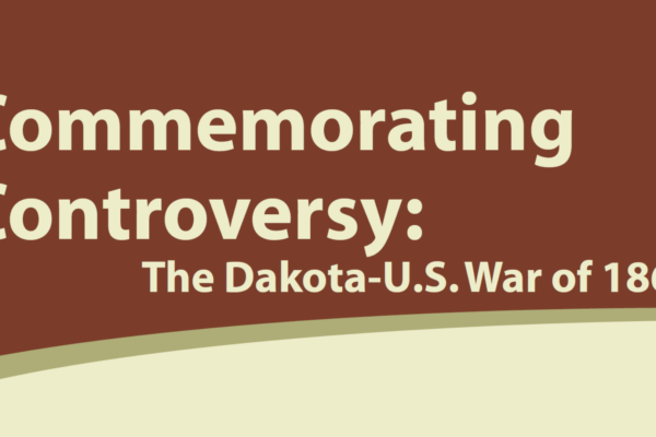 A sign reads: “Commemorating Controversy: The Dakota-U.S. War of 1862” in large, bold text on a brown and cream background with a curved border at the bottom.