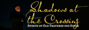 A person stands in the dark with arms crossed, wearing a hat and glasses. Large yellow script text reads, “Shadows at the Crossing.” Below, white text says, “Spirits of Old Traverse des Sioux.”.