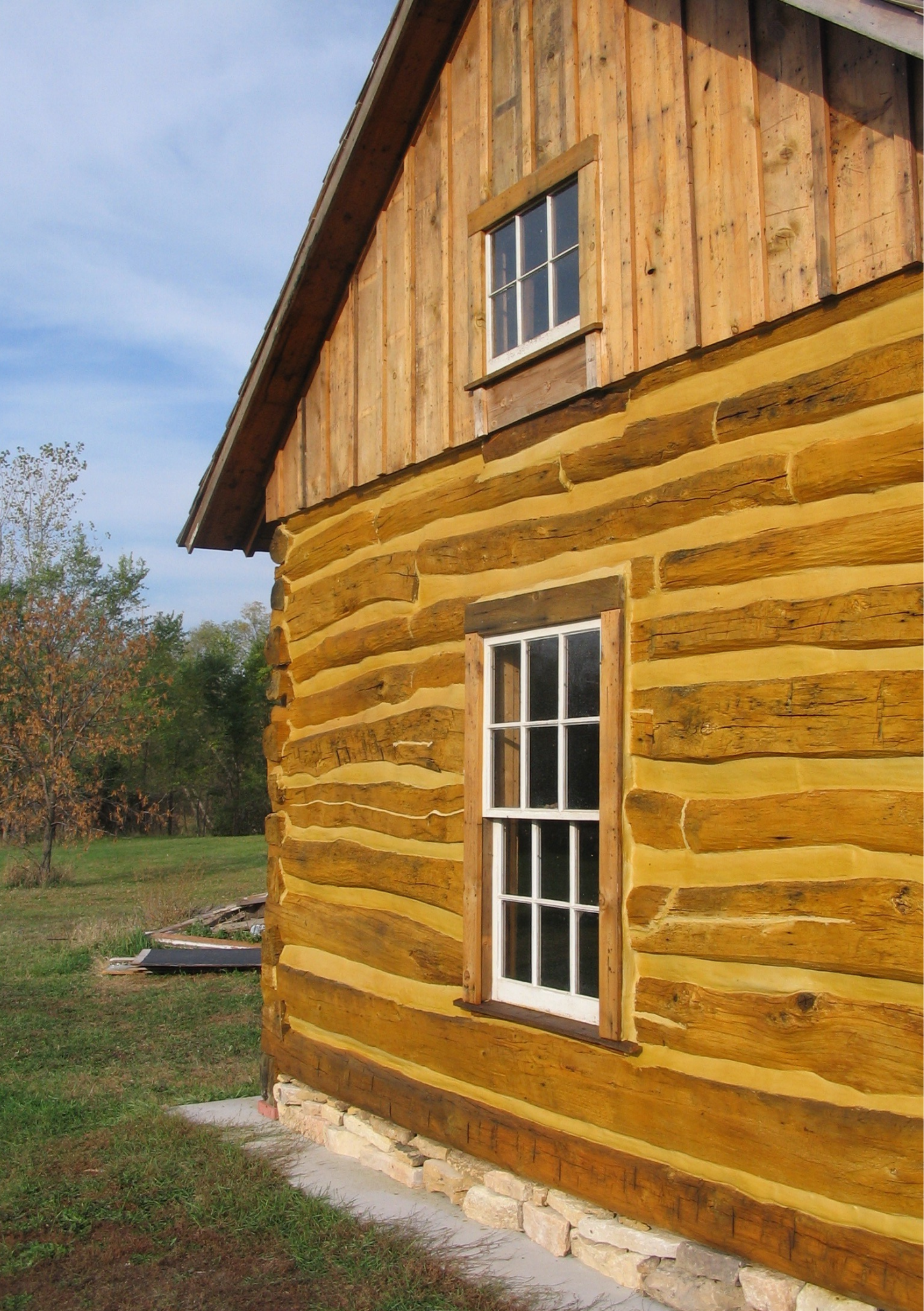 A side view of a log cabin with a stone foundation and two windows.